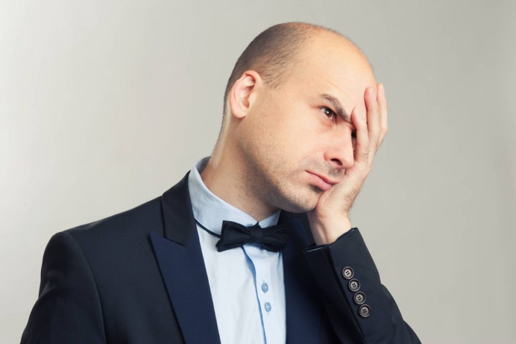 A bald man in a suit and bow tie holding his face with one hand, looking frustrated