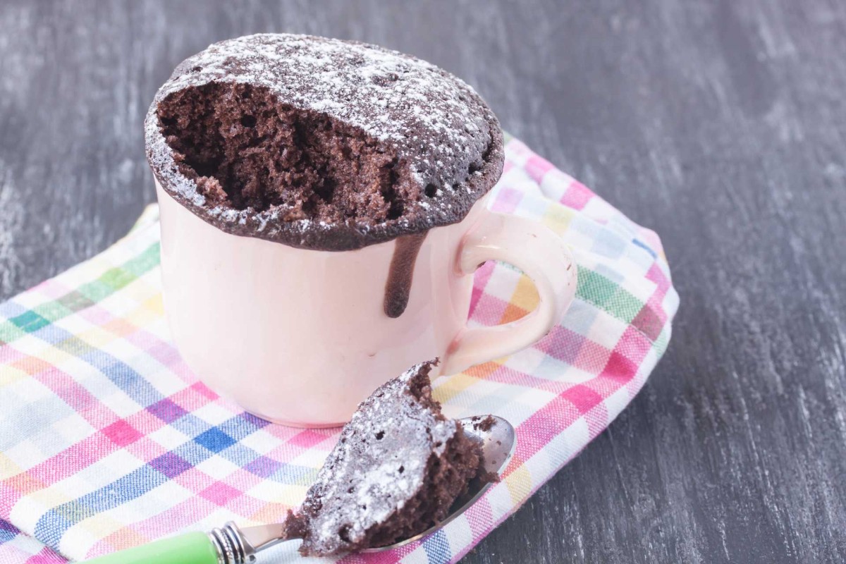 A chocolate mug cake overflowing from a mug, with a spoonful of cake beside it on a checkered cloth