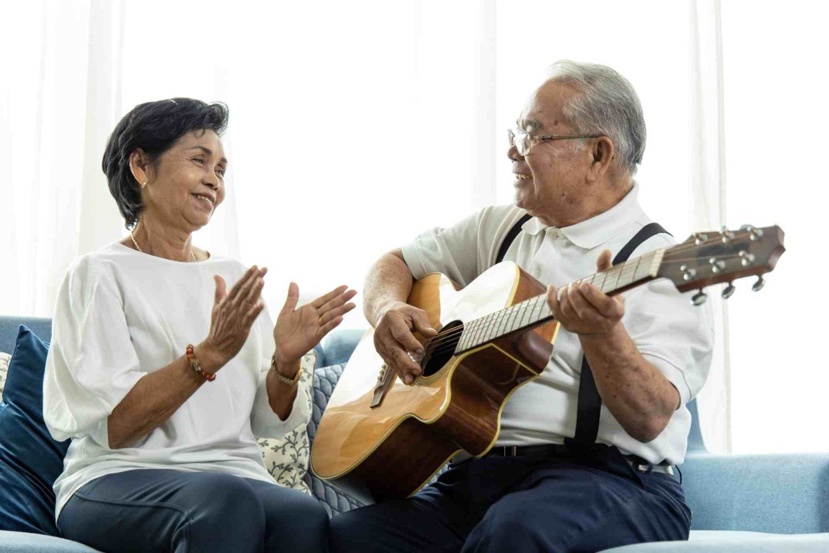 Elderly couple smiling at each other while playing guitar together at home