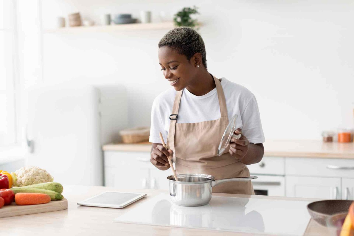 A woman wearing an apron smiles while stirring a pot on a stovetop in a clean, bright kitchen with vegetables nearby