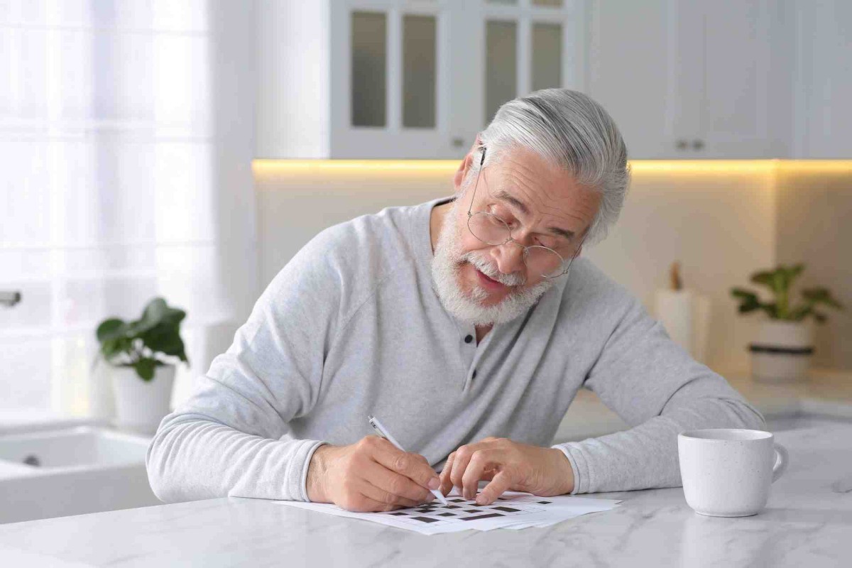 An older man with glasses working on a crossword puzzle at a table with a cup of coffee
