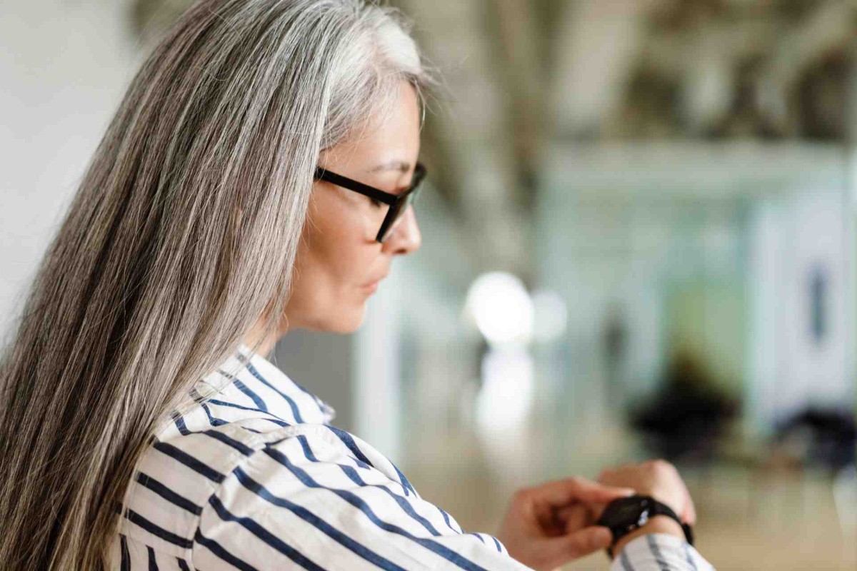 A woman with long gray hair and glasses looks at her smartwatch indoors